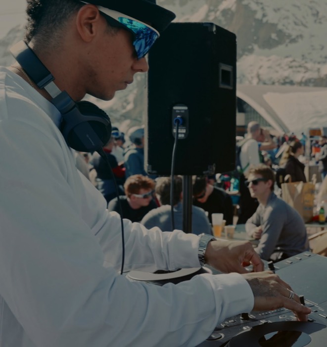 A DJ in sunglasses and headphones mixing outdoors, with skiers and snowy peaks behind