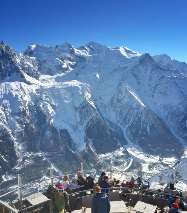 People at tables on a mountain terrace, the snow-covered Mont-Blanc massif behind under a deep blue sky